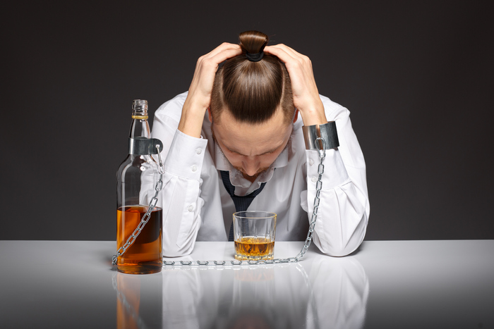 Na zdjęciu załamany człowiek trzyma głowę w dłoniach, obok stoi butelka alkoholu i szklanka. Jego dłoń i butelkę łączy łańcuch